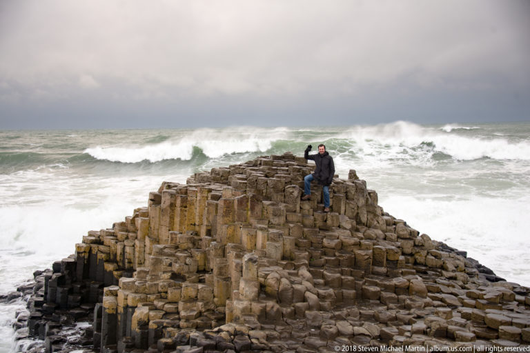 Visitor raising one arm from the top of a basalt mound as a large wave breaks behind.