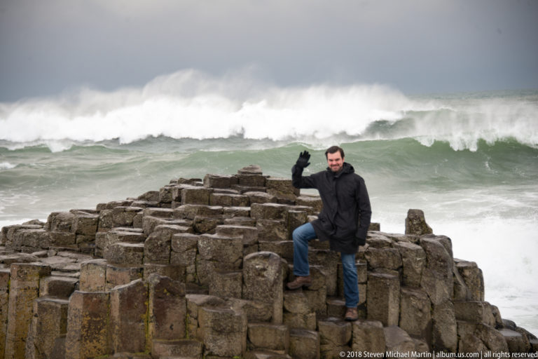 Visitor standing among tall basalt columns with one foot raised and a large breaking wave behind.