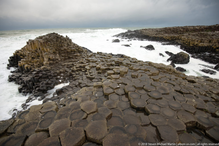 Wide wet basalt pavement with polygonal column tops leading toward a raised mound beside white surf.