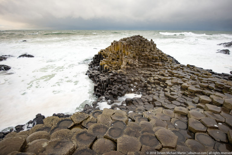 Elevated view of polygonal basalt extending toward a raised mound nearly surrounded by white Atlantic surf.