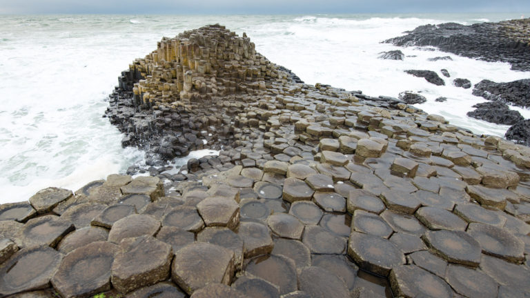 Large wet polygonal basalt blocks leading toward a steep mound of columns surrounded by white surf.