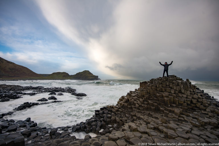 Visitor with raised arms atop a basalt mound, framed by surf, dark rock shelves, and storm clouds.