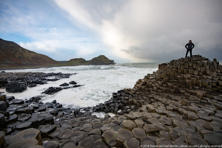 Visitor standing atop a stepped basalt mound beside a broad view of surf, dark rocks, and distant headlands.