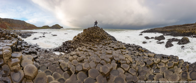 Long polygonal basalt ridge leading to a raised mound with a small visitor at its crest and surf on both sides.