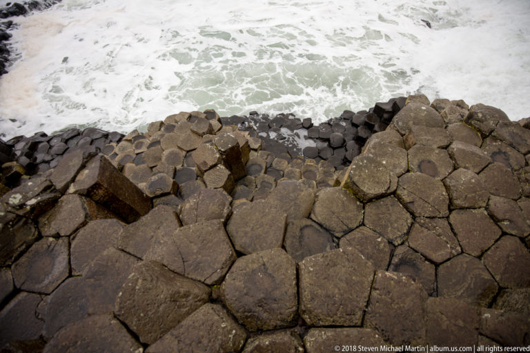 Downward view of wet polygonal basalt column tops ending above pale green water and white foam.