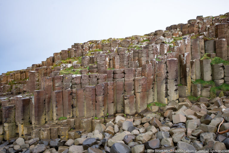 Wall of tall vertical basalt columns with broken blocks below and patches of green vegetation above.