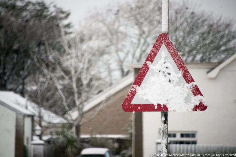 Triangular warning sign coated with snow, with only its red border visible beside houses and frosted trees.