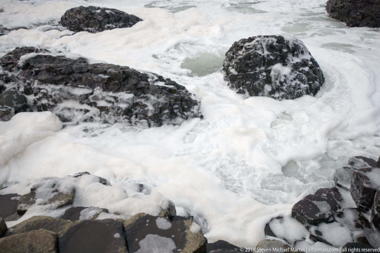 White foaming seawater swirling around dark basalt boulders along the Giant’s Causeway shore.