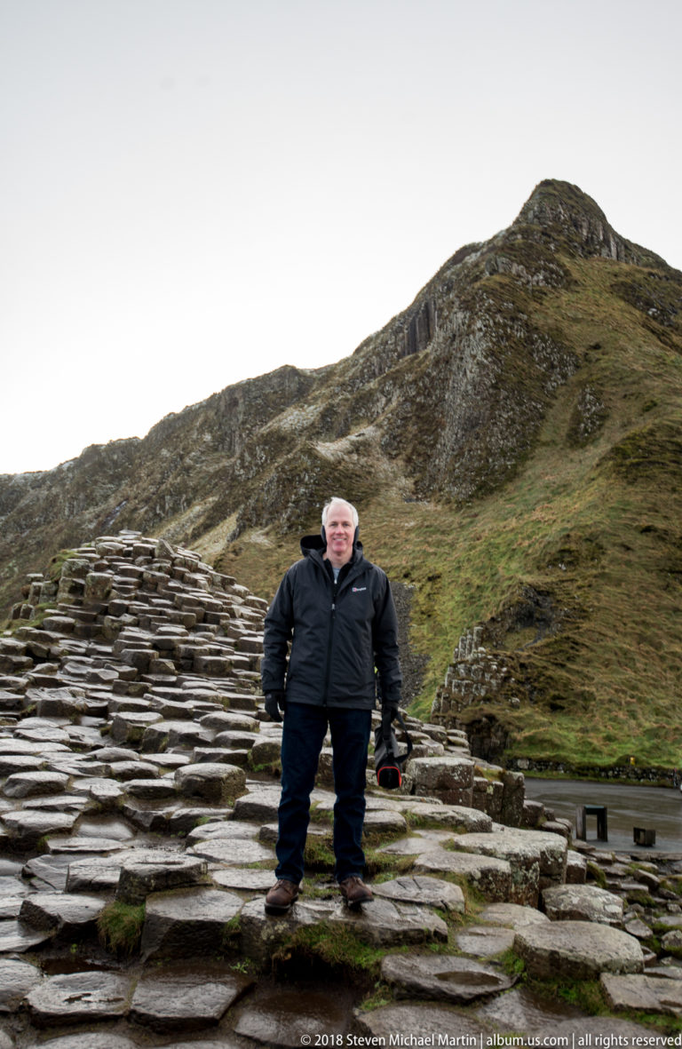 Visitor standing on wet basalt pavement with a steep grass-covered rocky cliff rising behind.