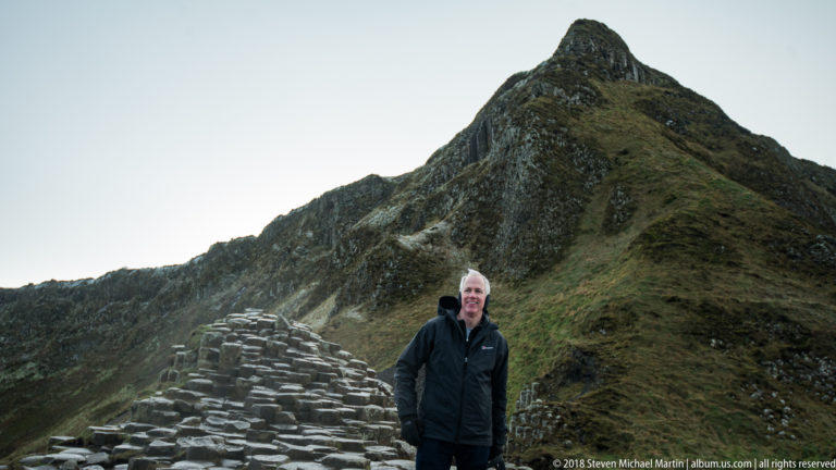 Visitor beside a low basalt mound beneath a steep pointed grass-and-rock coastal slope.