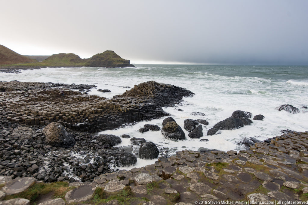 Basalt terraces and dark rocks separated by white surf, with a low green headland beyond.