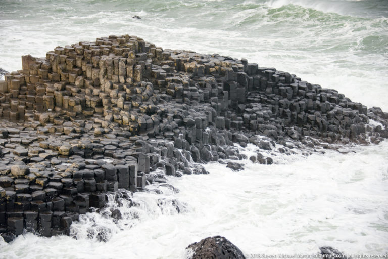 Terrace of tightly packed basalt columns descending into white surf, with taller ochre prisms at left.