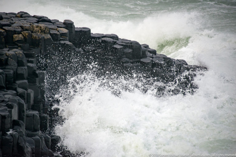 White Atlantic wave crashing against dark vertical basalt columns and throwing spray above the rock face.