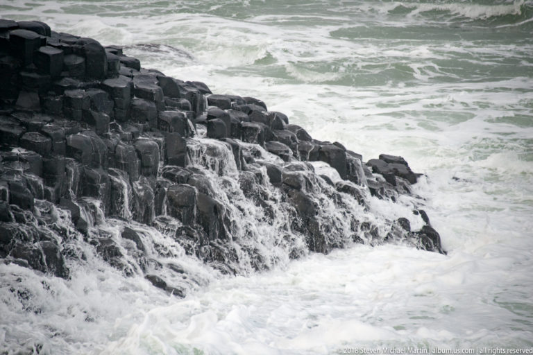 Seawater cascading in thin streams over stepped dark basalt columns into white foam below.
