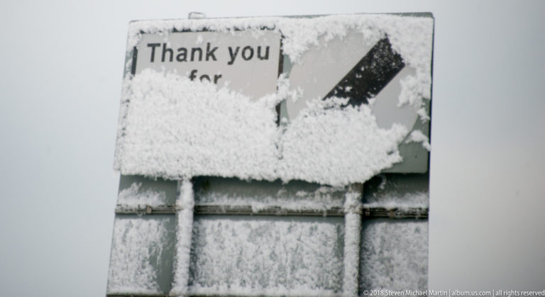 Snow and ice obscure most of a small directional sign, leaving a black right-pointing arrow and partial lettering visible.