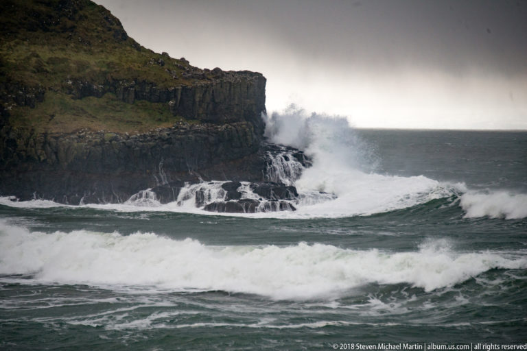 Large Atlantic waves breaking against a dark layered cliff with a grass-covered top and white cascades at its base.