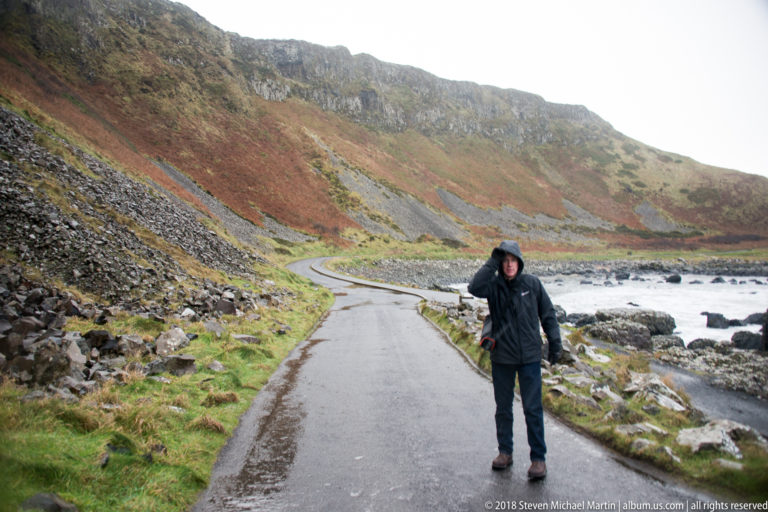 Visitor walking on a narrow coastal road between steep rocky slopes and the wave-washed shoreline.