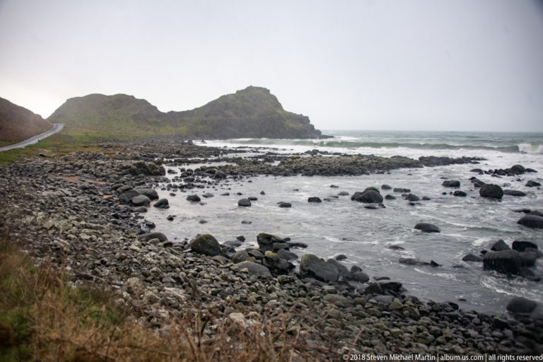 Rocky inlet with scattered dark boulders, rough gray-green water, and a low grass-covered headland.