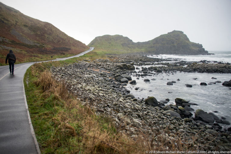 Visitor walking along a paved coastal route beside a rocky shore toward a low headland.
