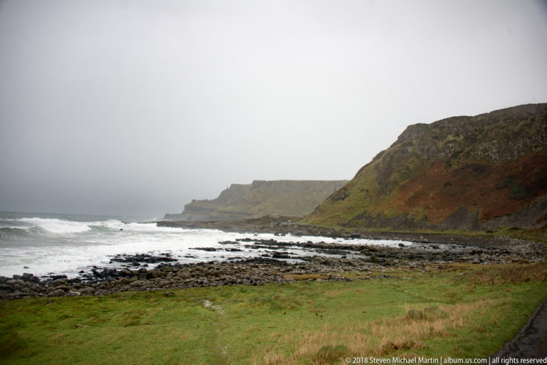 Stormy rocky bay with white surf between steep grassy headlands beneath a low gray sky.
