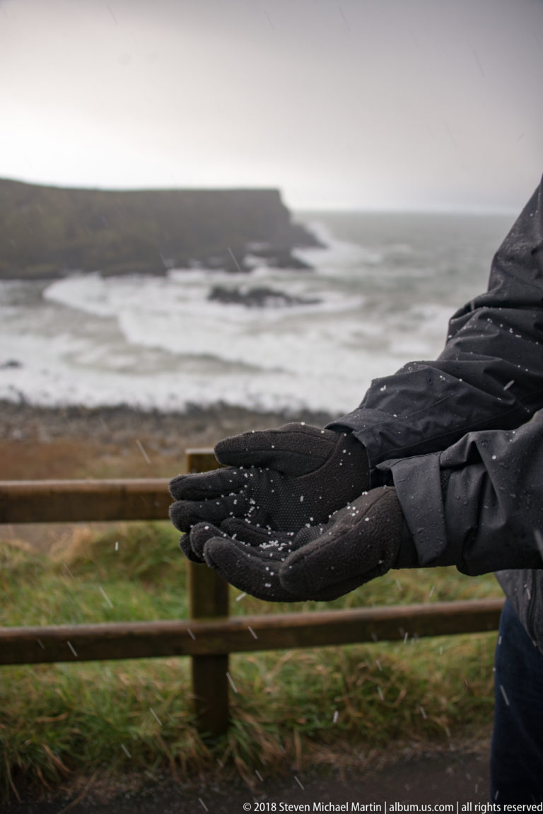 Black gloved hands holding scattered white hail or graupel pellets with the stormy coast blurred behind.