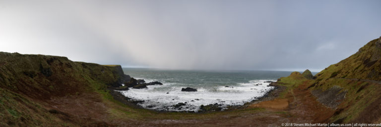 Rocky cove between steep grassy slopes with white surf and a dark curtain of precipitation offshore.
