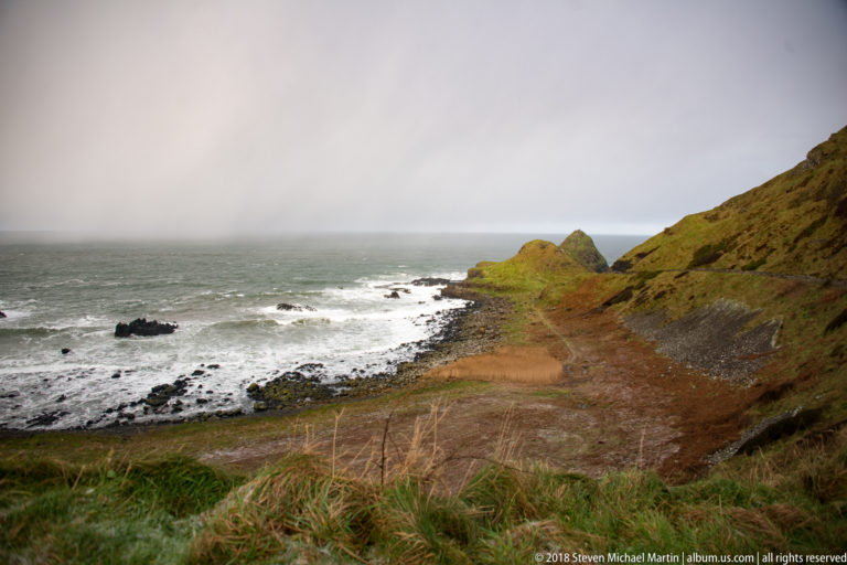 Green coastal headland above a dark rocky shore and white surf, with wintry precipitation on foreground grass.