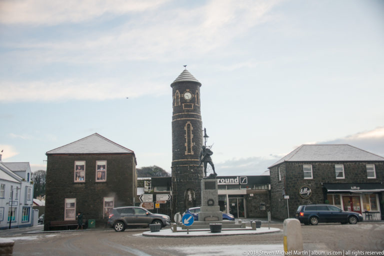 Dark stone clock tower at a village junction, surrounded by low buildings, parked cars, and patches of snow.