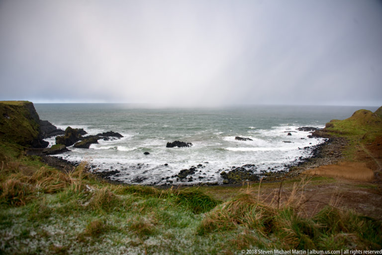 Elevated view of a rocky bay with white surf, dark outcrops, grassy headlands, and frosted foreground grass.
