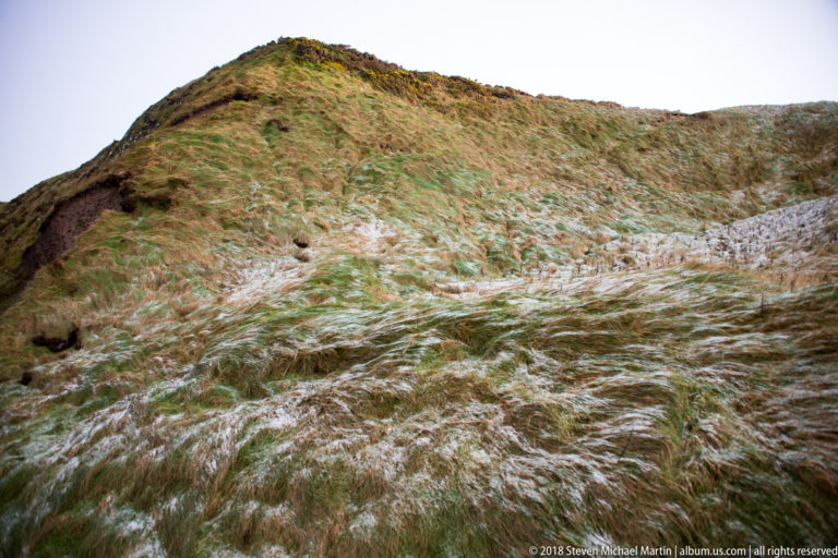 Steep slope covered in wind-swept grass with patches of white frozen precipitation and exposed dark earth.