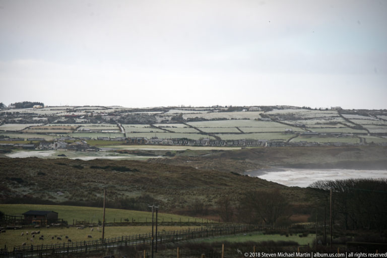 Patchwork coastal fields divided by hedgerows above a distant curved shoreline and pale surf.
