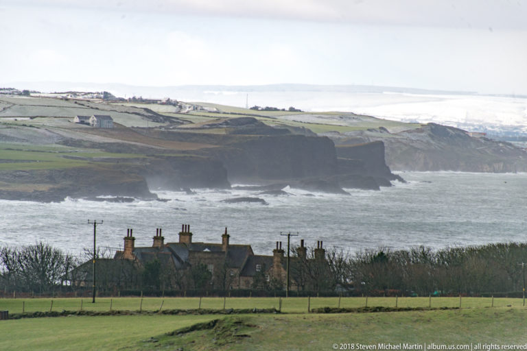 Rural houses and fields in front of dark coastal cliffs with white surf and the sea beyond.