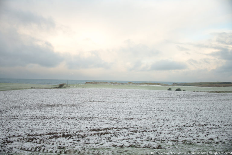Snow-dusted agricultural field stretching toward a low Atlantic coastline beneath broken winter clouds.