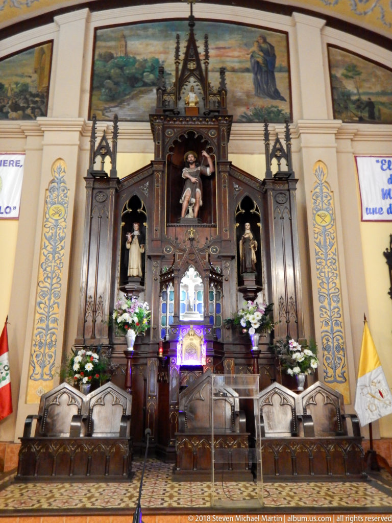 Dark carved church altar with statues, flowers, painted wall decoration, and ceremonial chairs across the sanctuary.