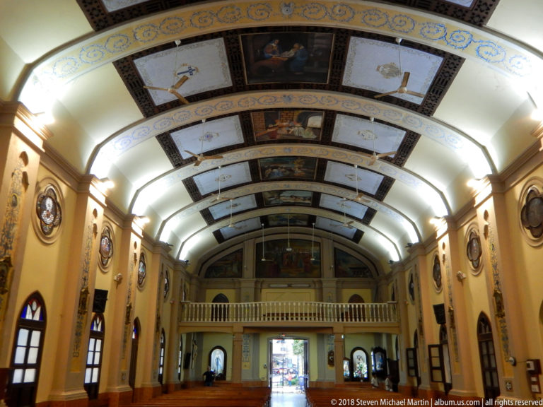 Long church nave with wooden pews, painted barrel-vault ceiling, stained-glass medallions, and white gallery.