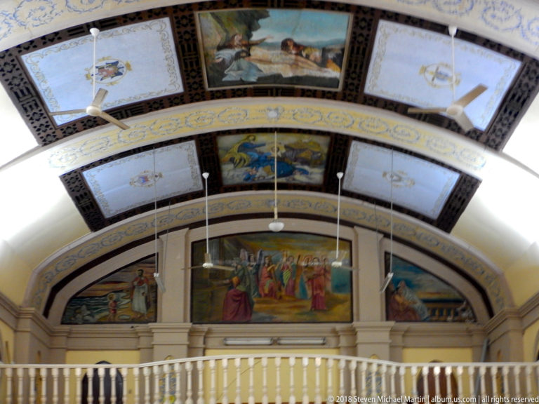 Painted barrel-vault church ceiling with religious panels above a white balustraded gallery and large mural.