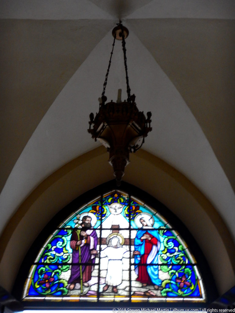 Dark hanging church lamp suspended above a glowing arched Holy Family stained-glass window.