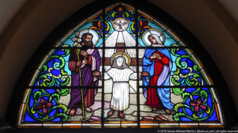 Arched stained-glass window of Joseph, Mary, and the child Jesus beneath a radiant heavenly figure.