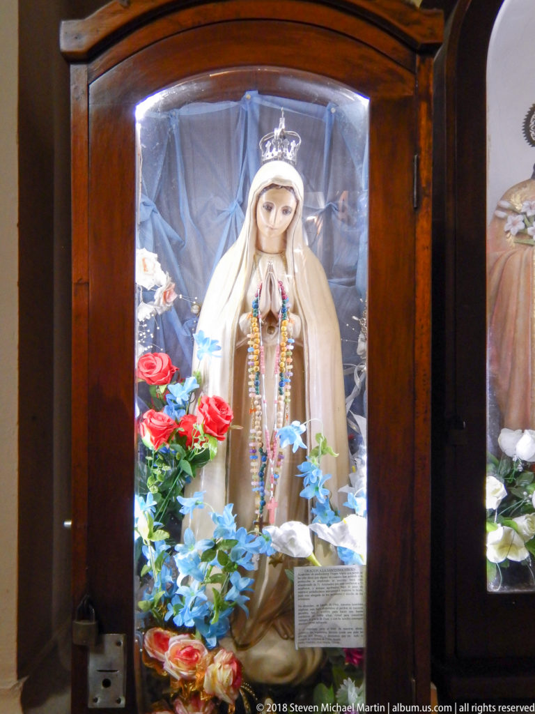 Crowned Virgin Mary statue with rosaries inside a wooden glass case surrounded by red, white, and blue flowers.