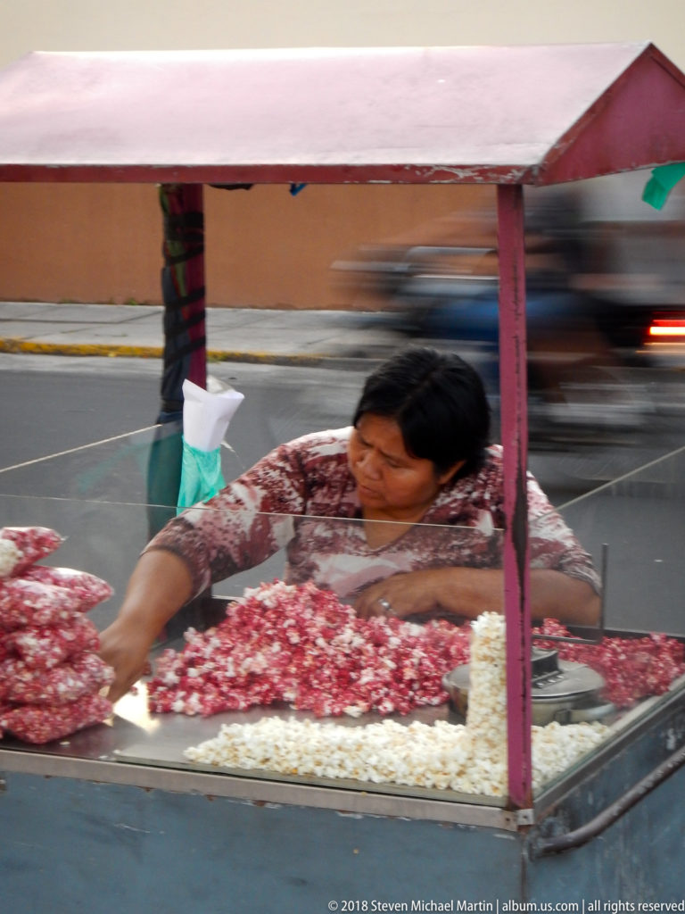 Woman reaching across a street cart filled with popcorn, with large bags of popcorn and blurred motorcycles behind.