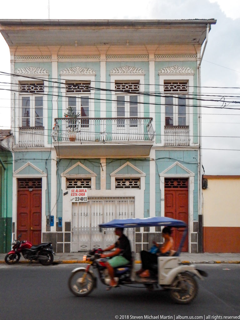 Mint-green and white two-story historic house with ornate windows, small iron balconies, red doors, and passing mototaxi.