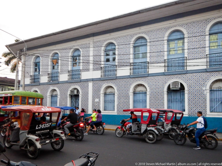 Large blue-and-white tiled two-story building with rows of arched windows, iron balconies, and mototaxis outside.