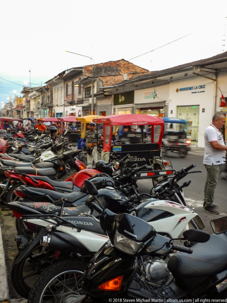 Rows of tightly parked motorcycles with red-canopied mototaxis and shops behind them in central Iquitos.
