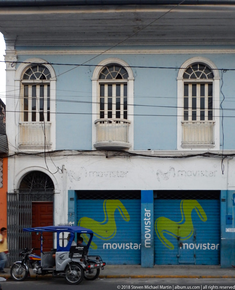 Blue-and-white historic upper façade with three arched balcony windows above blue Movistar storefront shutters.