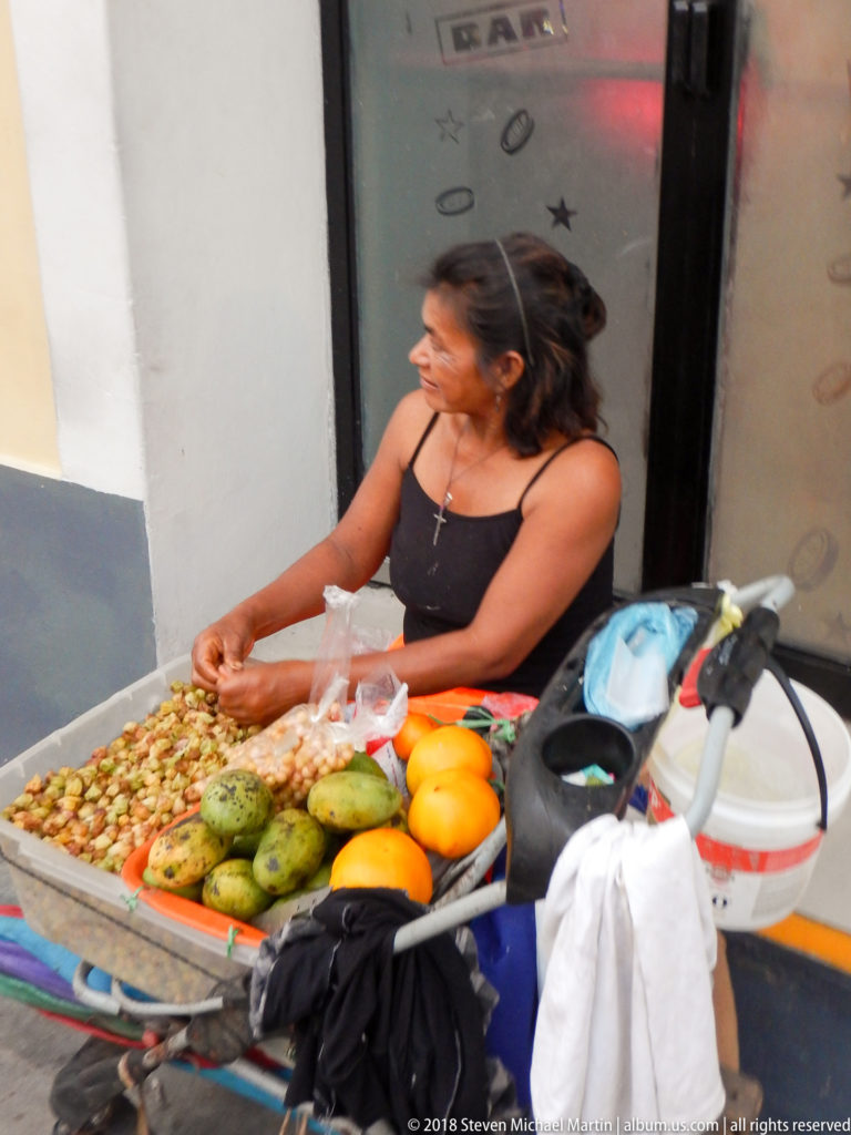 Woman seated at a sidewalk cart covered with green, yellow, and bagged small tropical fruits.