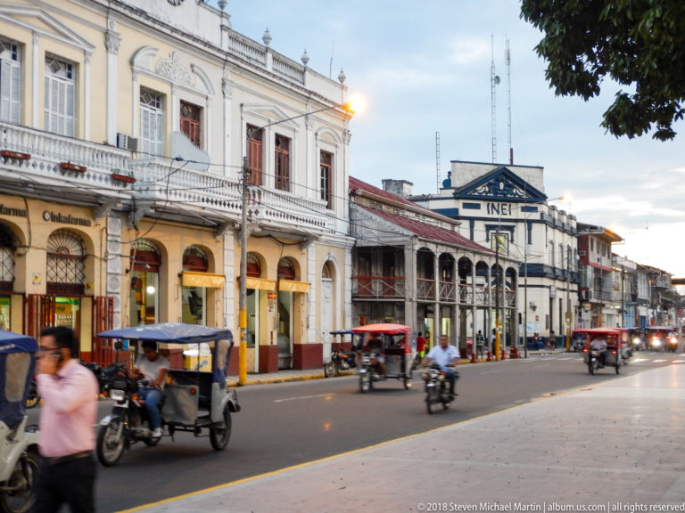 Historic arcaded buildings and INEI signage along a busy Iquitos street with mototaxis and motorcycles.
