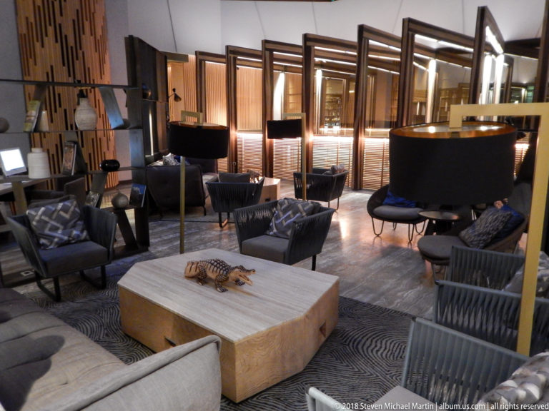 Contemporary hotel lounge with dark chairs, sofas, black lamps, wood screens, and a large pale coffee table.