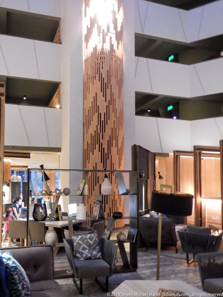 Modern hotel lobby with dark lounge seating beneath a tall illuminated vertical wood-slat feature.
