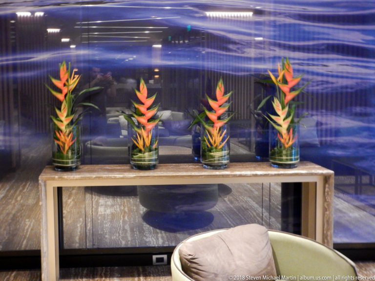 Four glass vases of orange heliconia arranged on a console against a reflective blue wall.