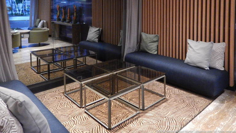 Dark-blue banquette with pale cushions, glass-and-metal tables, wood-slat wall, and patterned rug.